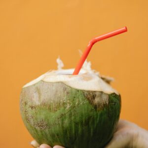 Home A hand holds a fresh coconut drink with a red straw against an orange background, exuding tropical vibes.